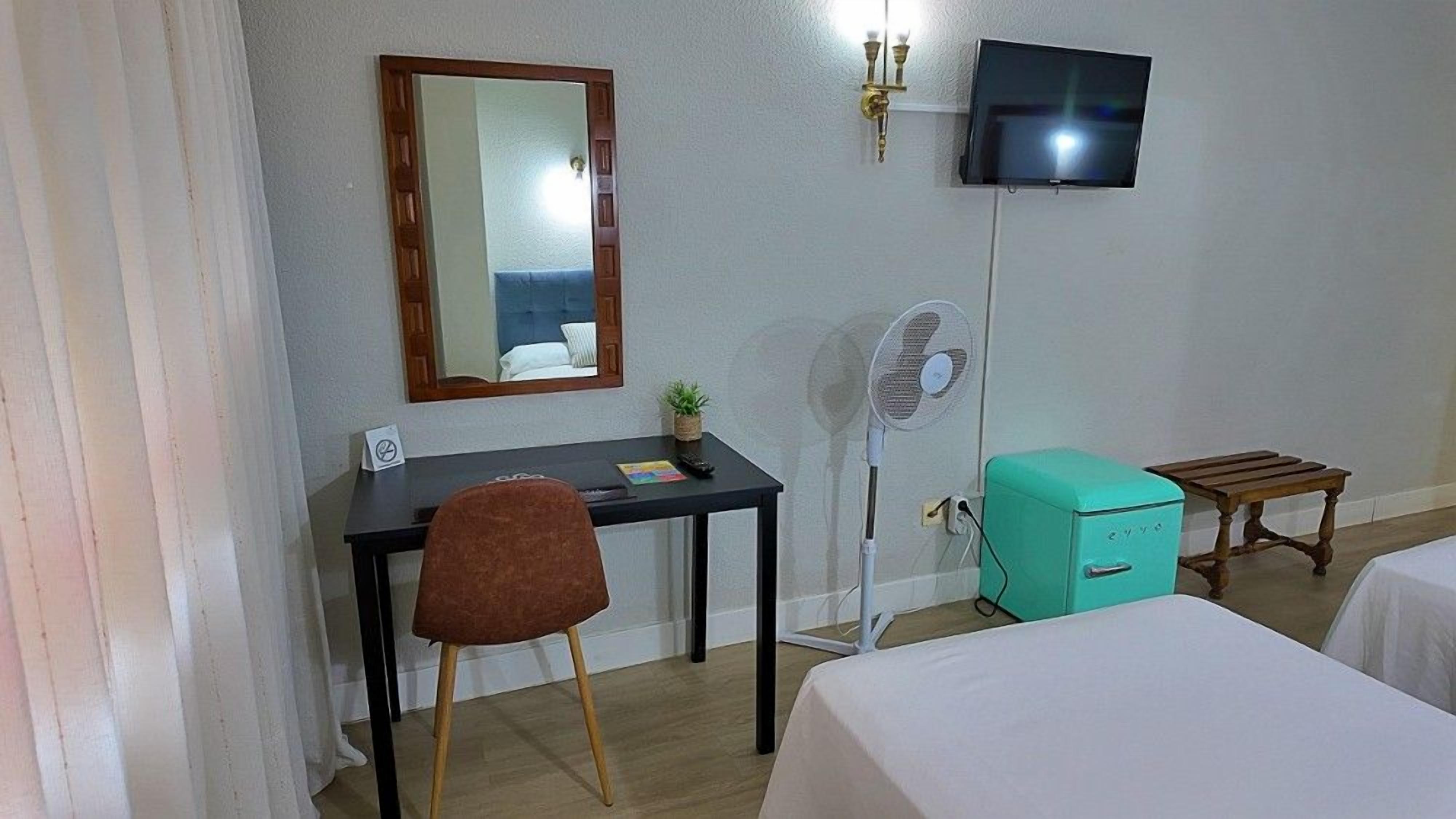 Una habitación de hotel con escritorio, silla, televisor, ventilador y dos camas.