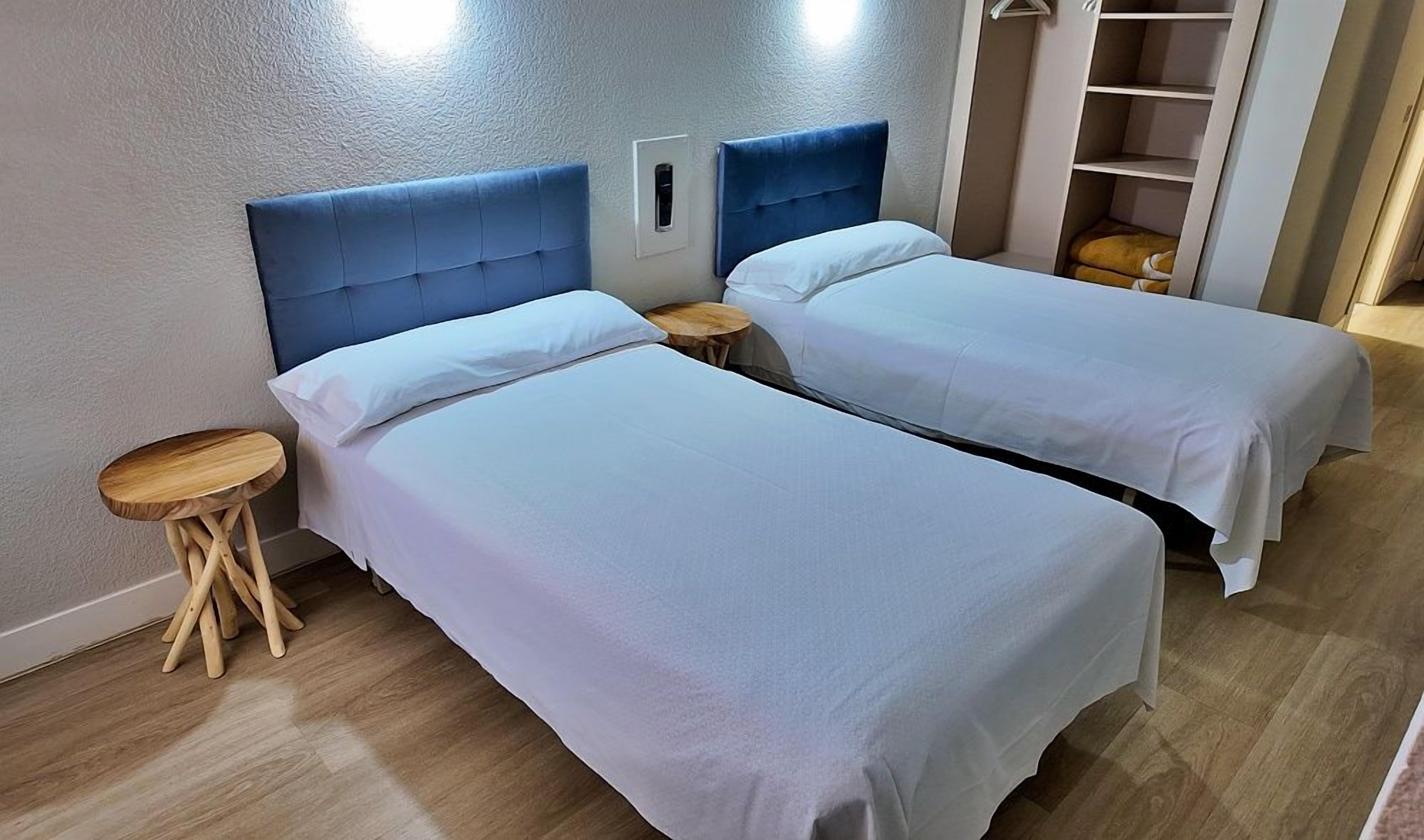Una habitación con dos camas, cada una con cabecera, un taburete de madera pequeño y una estantería al fondo.