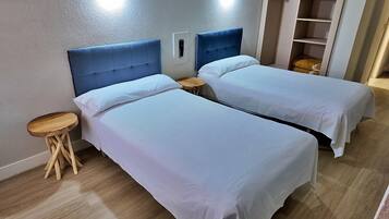 Una habitación con dos camas, cada una con cabecera, un taburete de madera pequeño y una estantería al fondo.