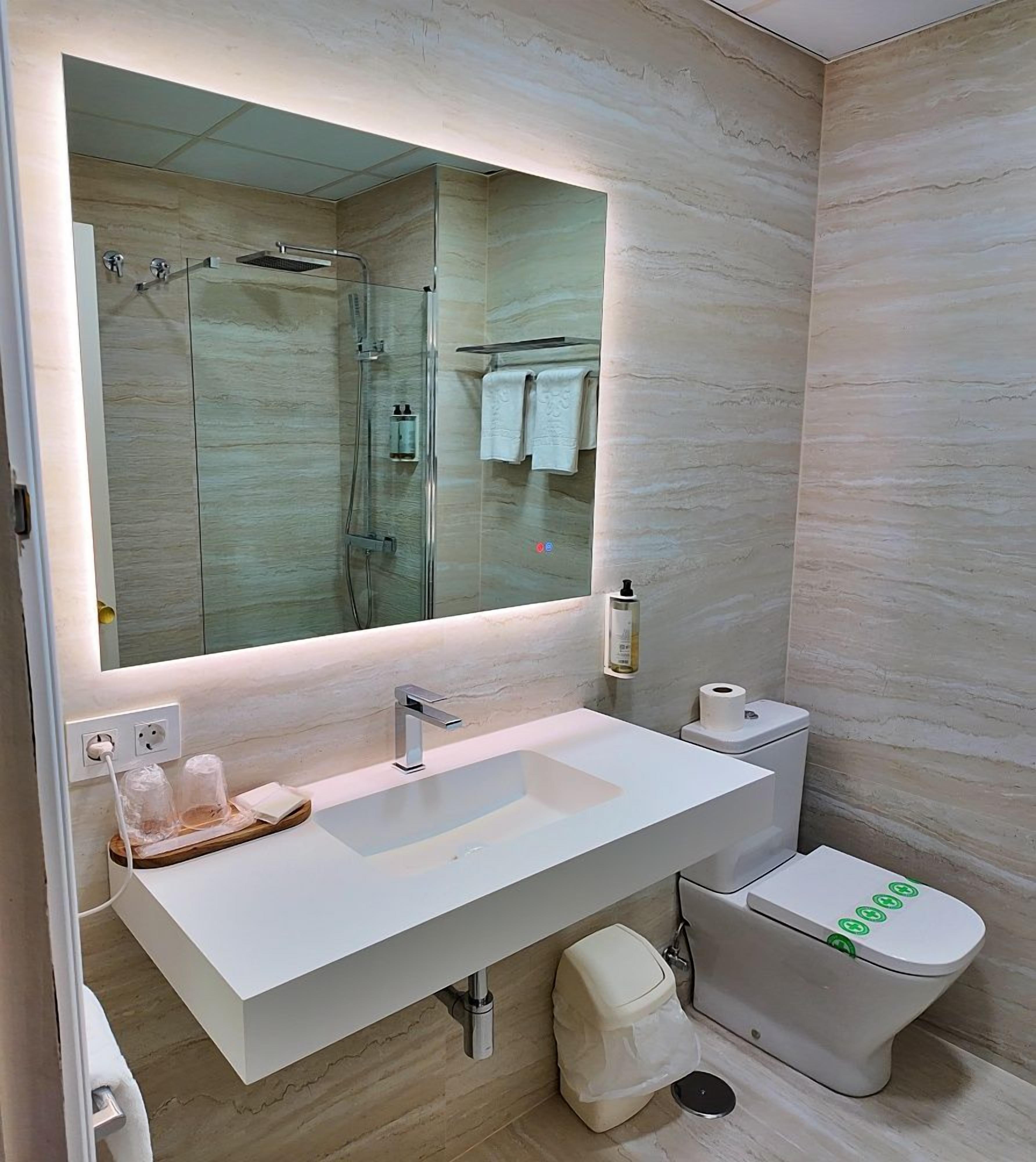 Un baño moderno con un espejo grande, un lavamanos blanco, un inodoro y una zona de ducha.