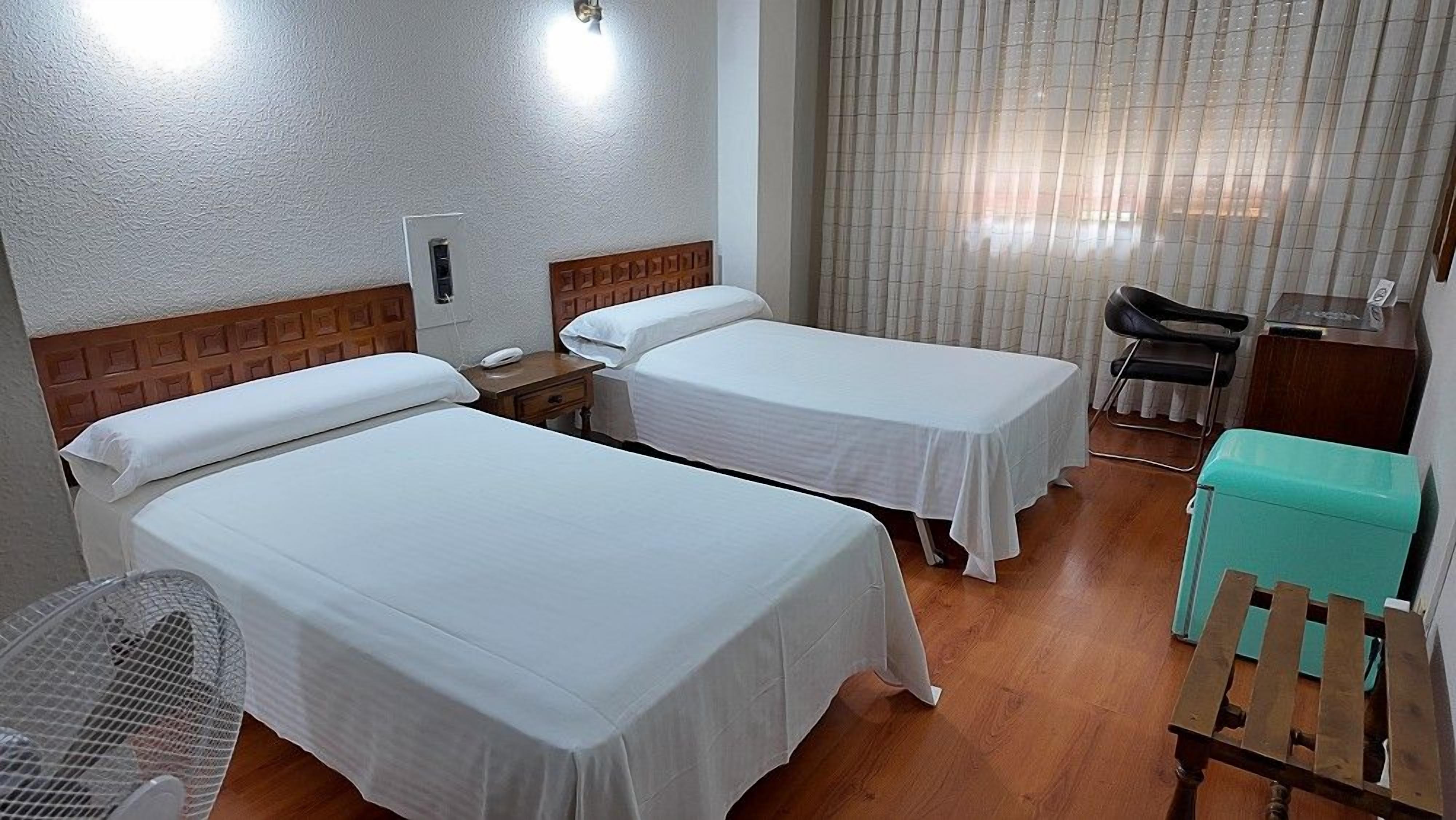 Una habitación de hotel con dos camas individuales, un escritorio, una silla, una mesita y un ventilador.