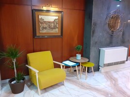 Uma poltrona amarela, uma pequena mesa redonda com uma planta em vaso, um quadro na parede e um radiador.