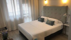 Una cama bien hecha con cabecera, lámpara de noche y ventana con cortinas de gasa.