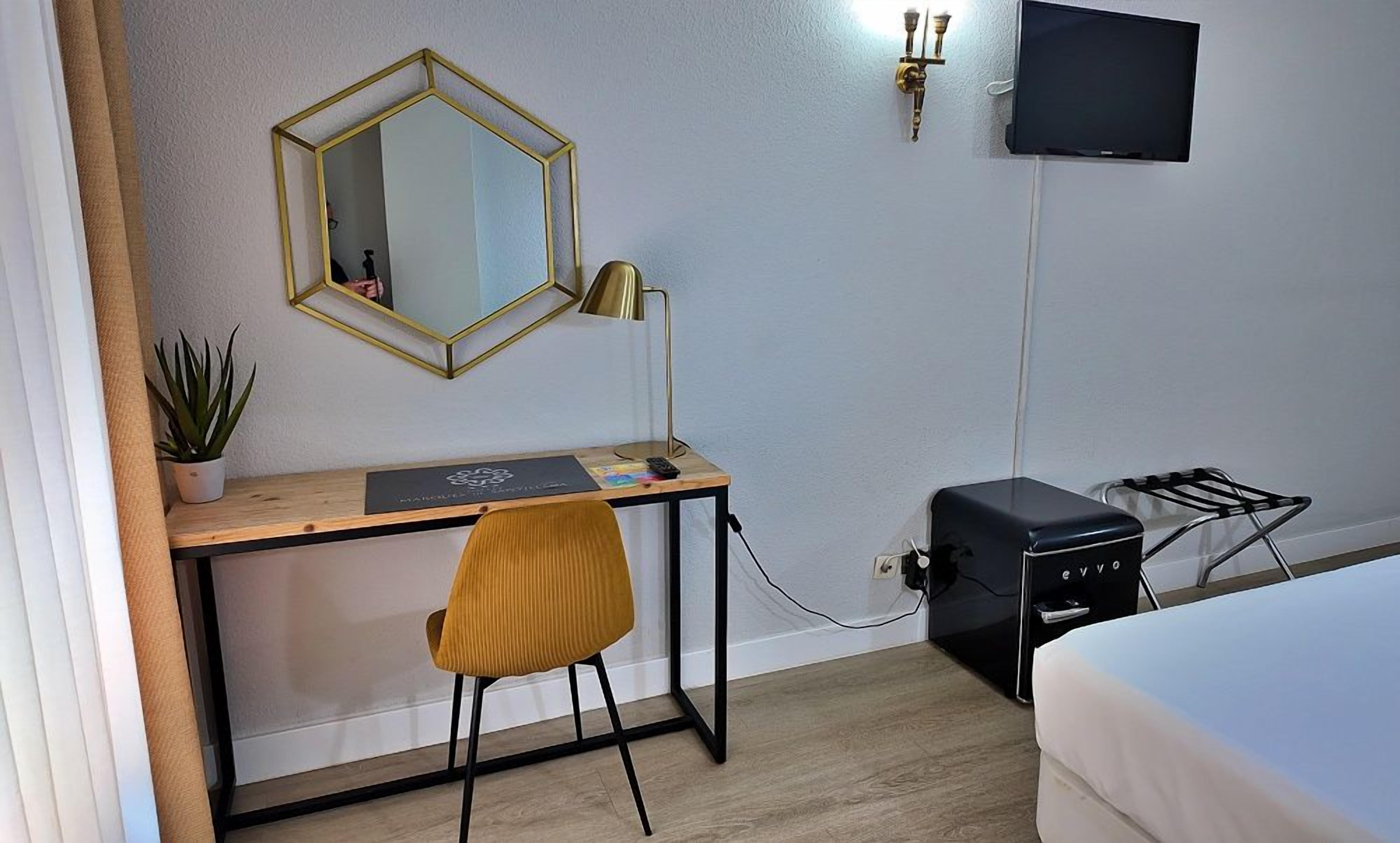 A hotel room with a wooden desk, a chair, a bed, a TV, and a charging station.