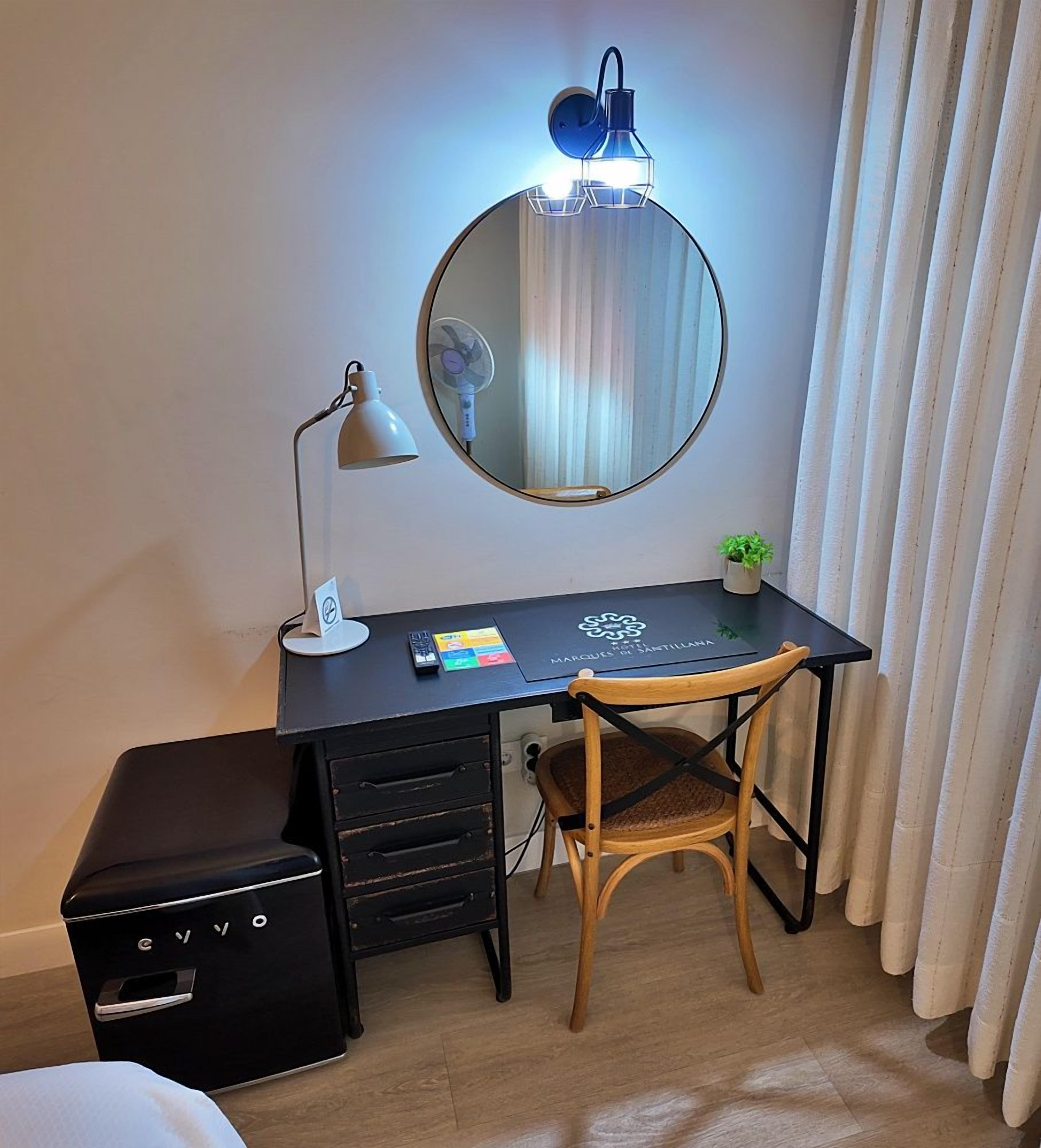 Habitación de hotel con escritorio, silla, espejo, lámpara, planta pequeña y maleta negra.