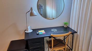Habitación de hotel con escritorio, silla, espejo, lámpara, planta pequeña y maleta negra.