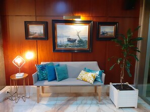 Una habitación con pared de madera, un sofá gris con cojines estampados, una mesita con una lámpara y una planta en maceta.