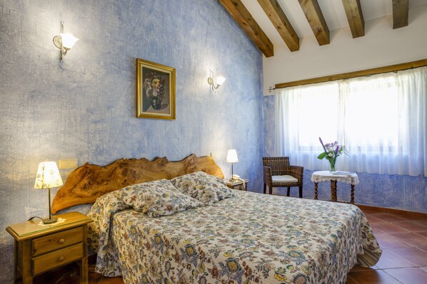 A bedroom with a wooden headboard, floral bedspread, bedside lamps, a small table with a vase, and a window with curtains. - Hotel Rural y SPA La Senda de los Caracoles (Ayllon)