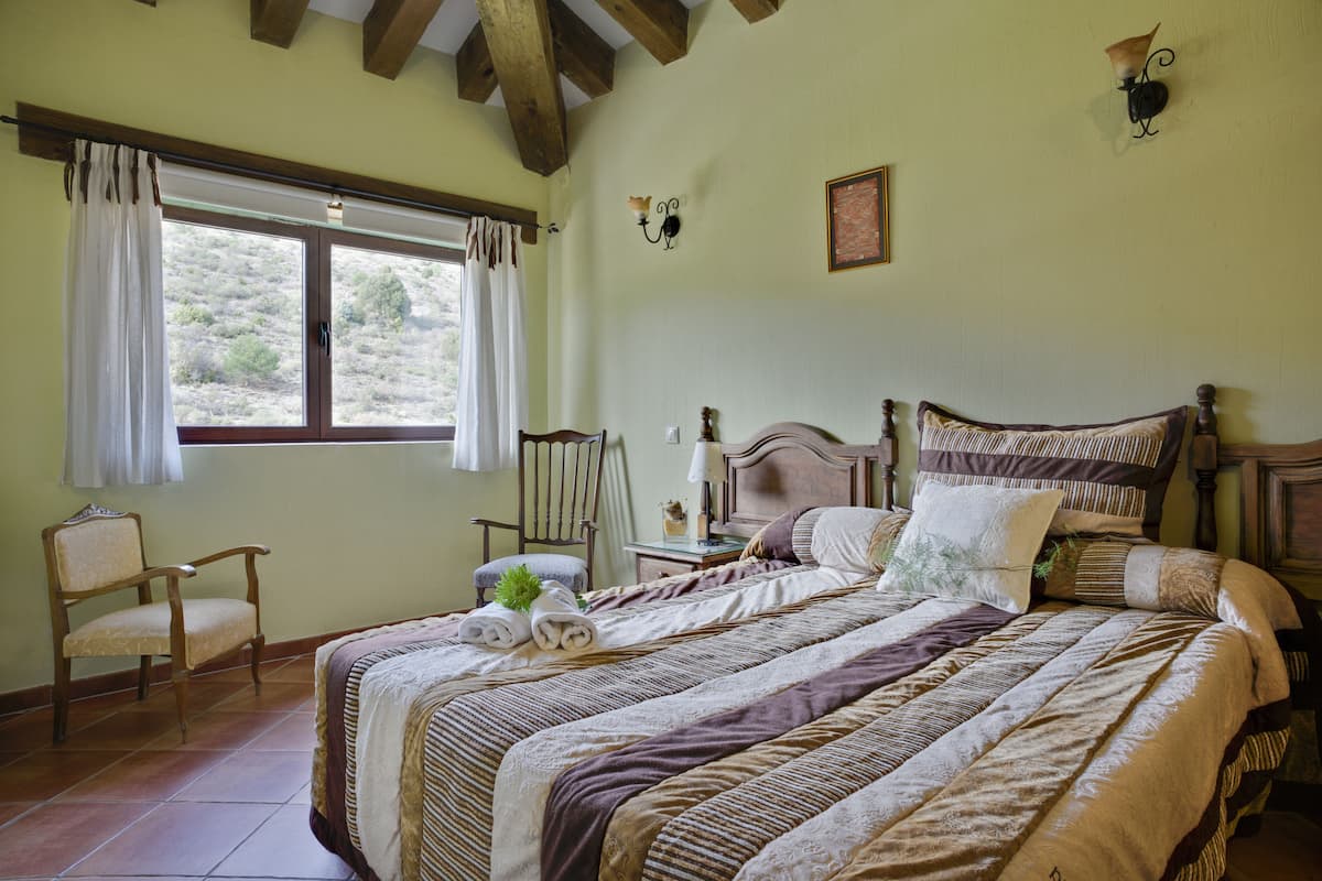 Ein Schlafzimmer mit einem großen Bett, einem Sessel, einem Nachttisch und einem Fenster mit Vorhängen.