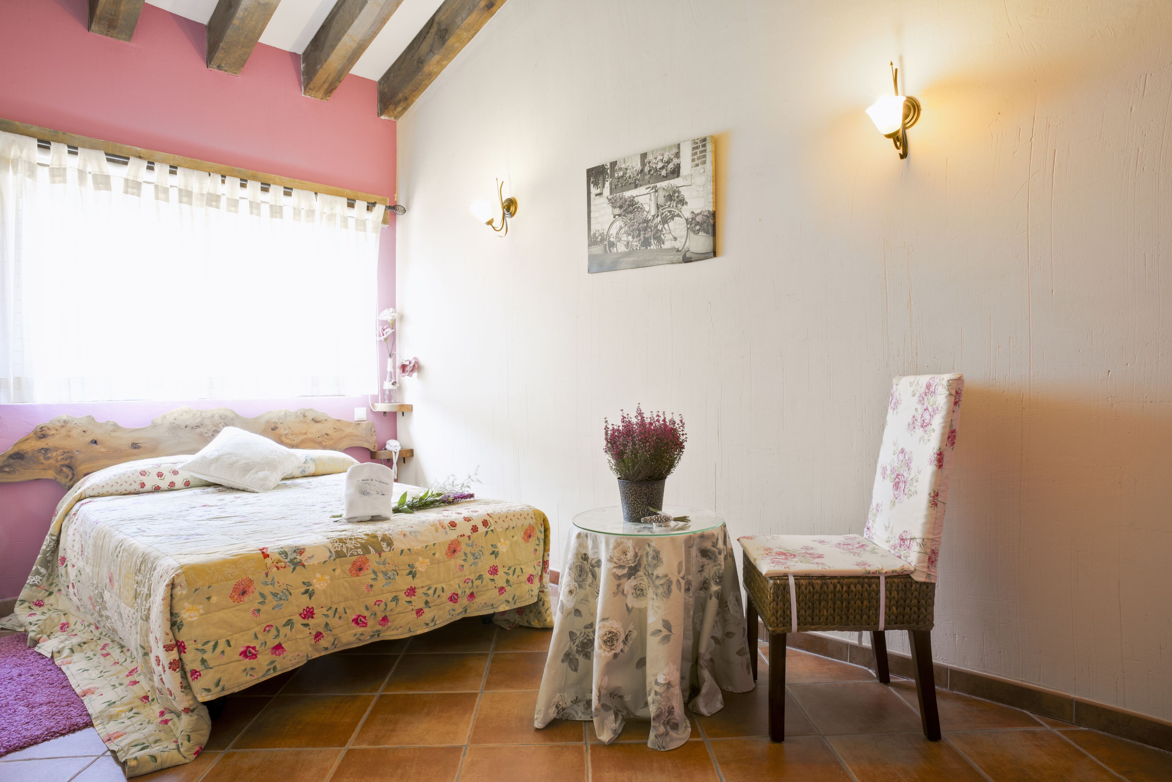 Un dormitorio con cama, una silla, una mesita con una flor y un cuadro colgado en la pared.