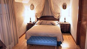 Un dormitorio con cama a dosel, suelo de madera, un banco azul y dos mesitas de noche con lámparas.