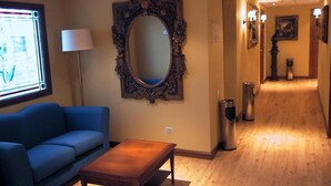 Un hall de hotel con un sofá azul, una mesa de centro de madera, un espejo grande y decorativo, y un letrero que indica la salida.