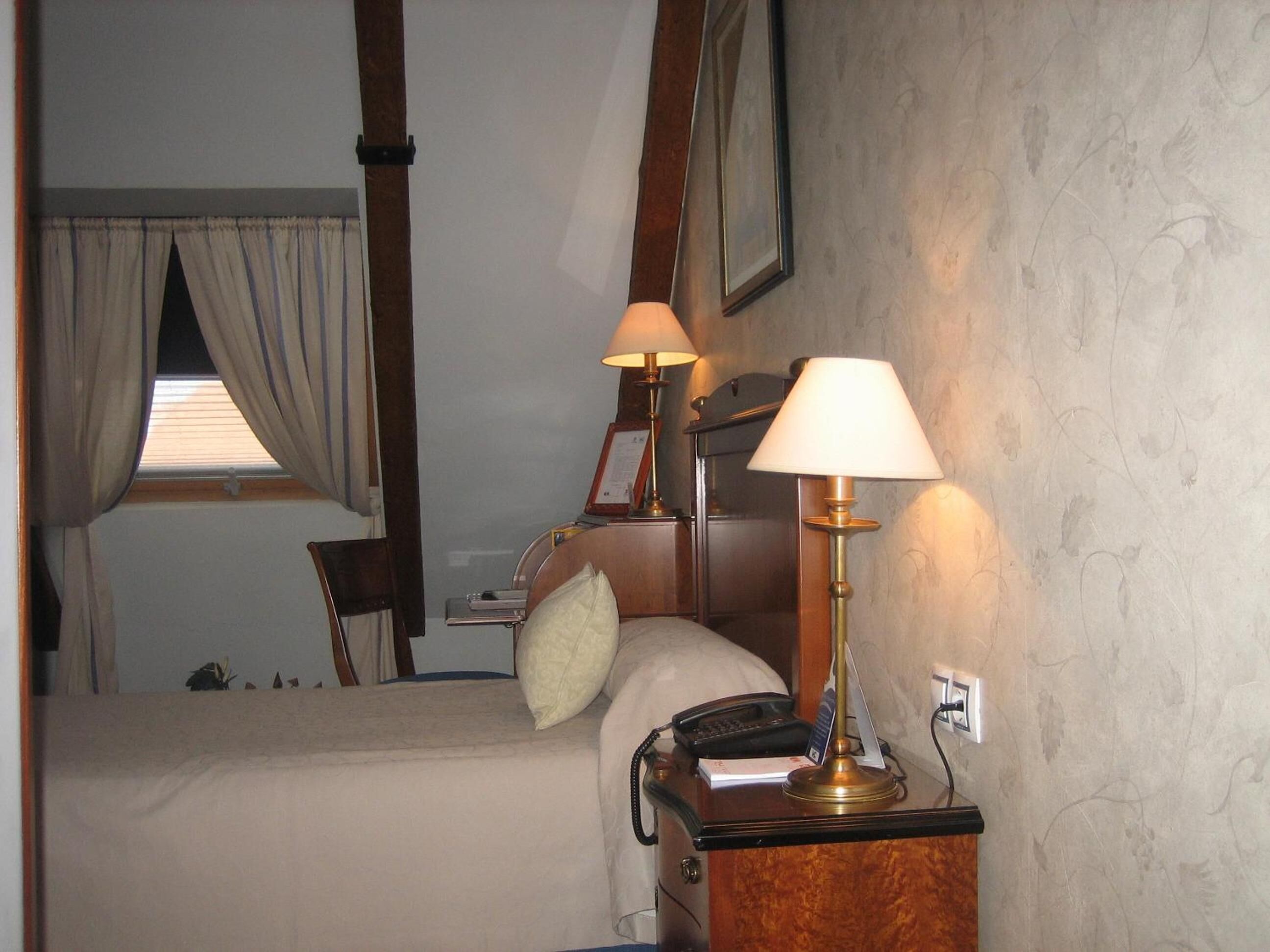 A hotel room with a bed, two bedside lamps, a nightstand with a phone and a lamp, a window with curtains, and a framed picture on the wall.