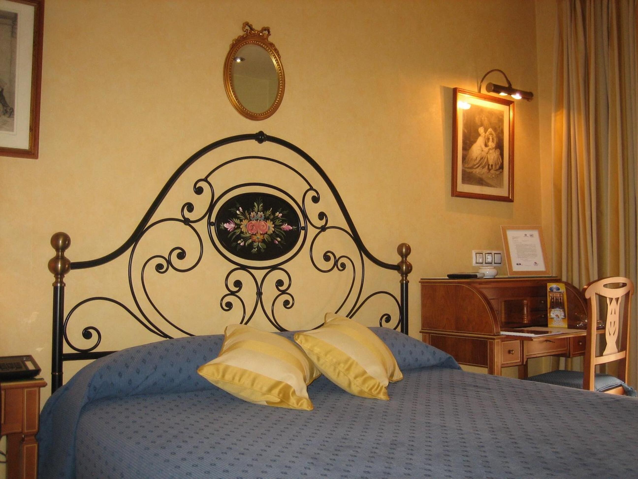 A bed with a headboard, a bedside table, a desk, a chair, a framed picture, and a wall-mounted mirror.