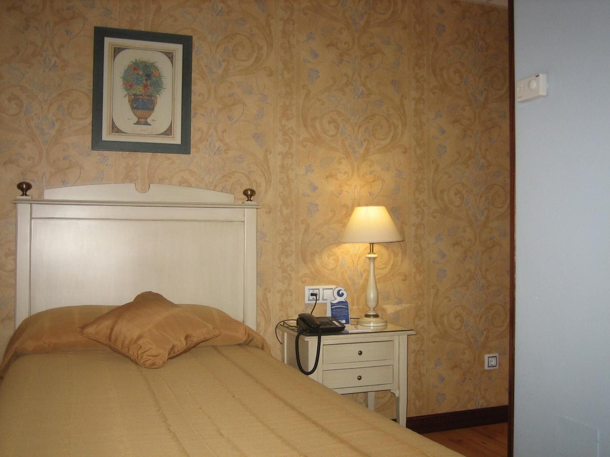Un lit bien fait, avec une couverture beige et des oreillers, une table de chevet avec une lampe, un téléphone et un tableau encadré au mur.