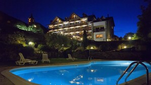 Ein Schwimmbecken mit Liegestühlen, ein Gebäude mit mehreren Balkonen und im Hintergrund ein Kirchturm.