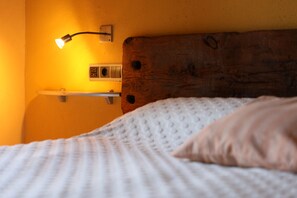 A bed with a white textured blanket and a single pillow, a wooden headboard, and a wall-mounted light fixture. - Font Del Genil (Arseguel)