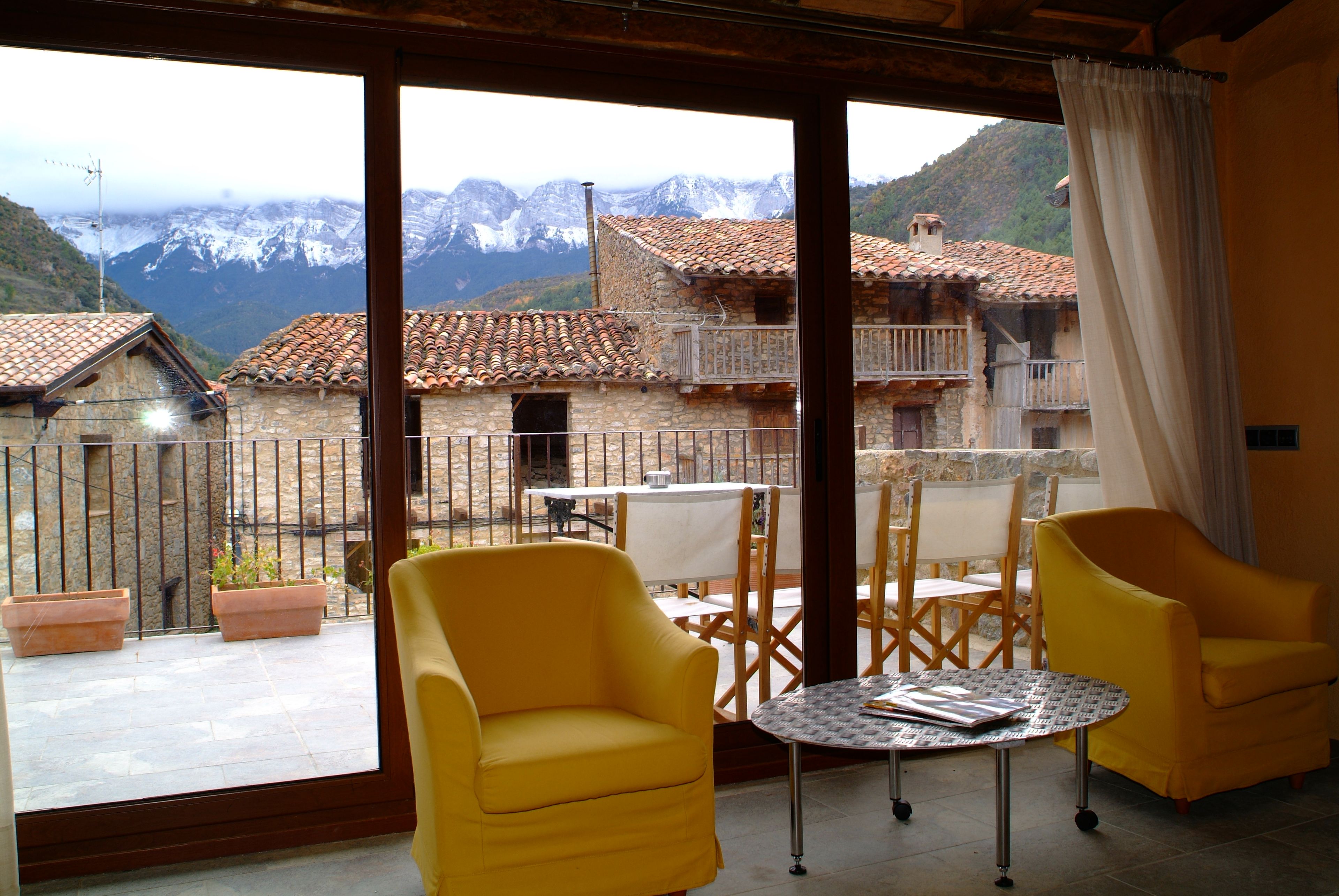 Un balcon avec des chaises jaunes et une table, d’où l’on peut admirer un paysage montagneux parsemé de bâtiments traditionnels.