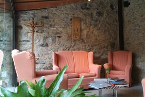 A room with stone walls, two pink armchairs, a wooden coat rack, and a small table with books and a vase. - Font Del Genil (Arseguel)