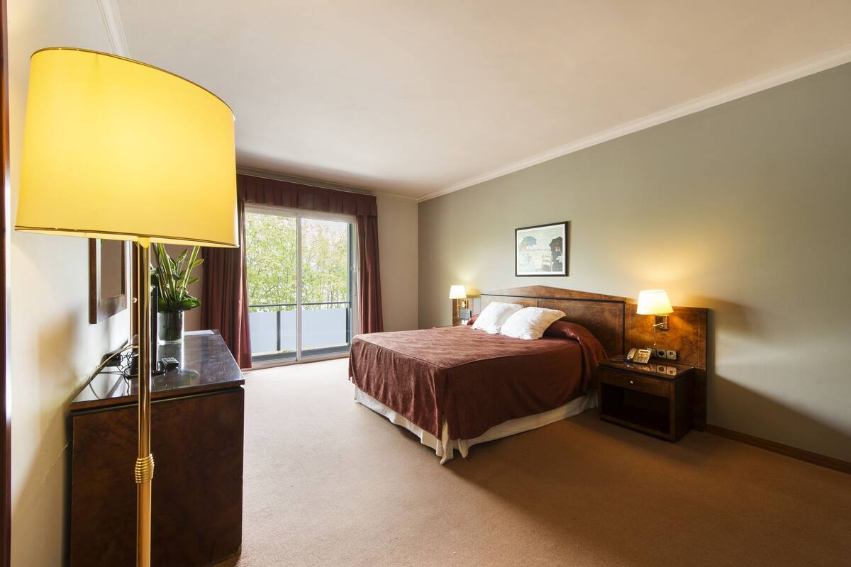A hotel room with a bed, bedside lamps, a desk with a lamp, a window with curtains, and a framed picture on the wall.