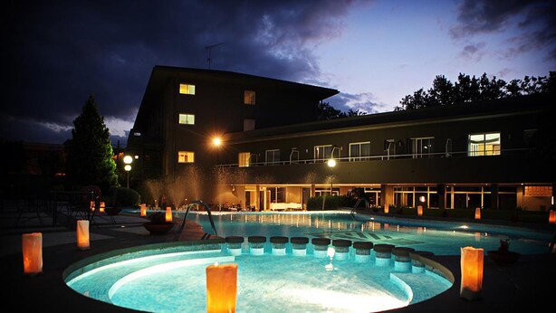 Une piscine avec un espace de jacuzzi circulaire, entouré de piliers illuminés par des bougies, et un bâtiment avec de nombreuses fenêtres et des balcons en arrière-plan.