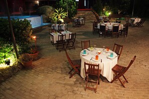 Un espacio de comedor al aire libre con mesas y sillas redondas, rodeado de plantas en macetas y una piscina.