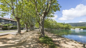 Une promenade au bord de la rivière, bordée d’arbres, un immeuble doté de balcons et une vue sur un lac et des montagnes au loin.