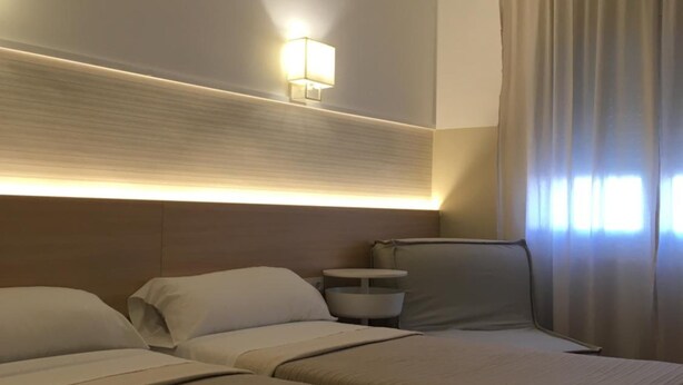 Habitación de hotel con dos camas, una mesita de noche, una lámpara y un ventanal con cortinas.