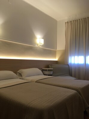 Quarto de hotel com duas camas, uma mesa de cabeceira, uma luminária e uma janela com cortinas.