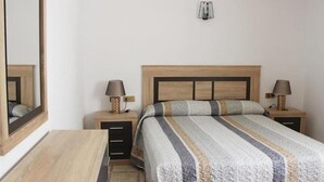 A bedroom with a wooden bed, two bedside tables with lamps, a mirror, and a small wall-mounted shelf. - Las Americas (Blanes)