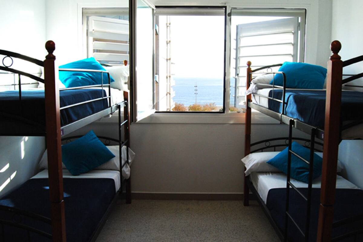 Een kamer met stapelbedden, een raam met zonwering en uitzicht op zee.