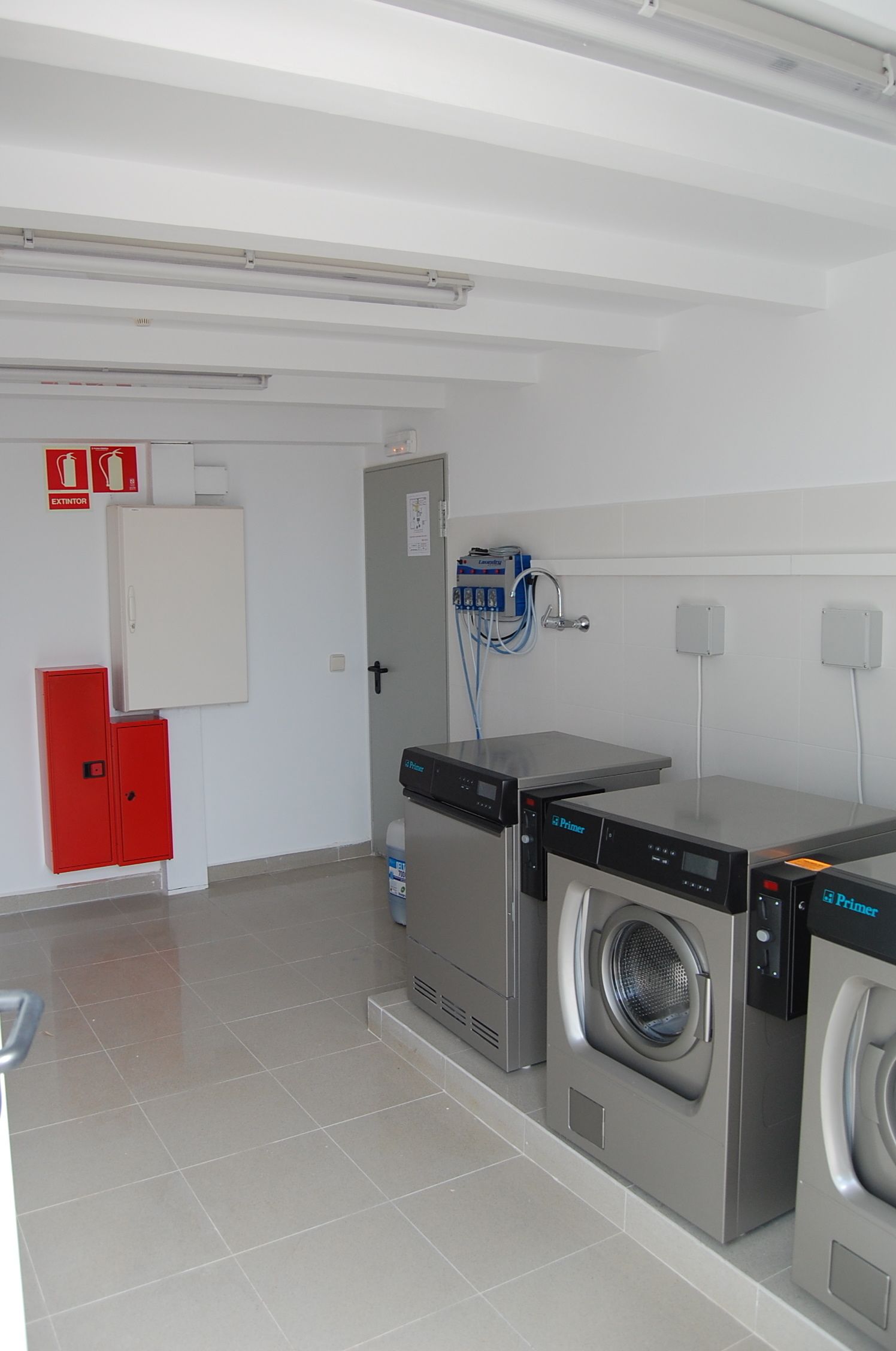 A laundry room with two front-loading washing machines, a red utility cabinet, and a door with a fire extinguisher sign.