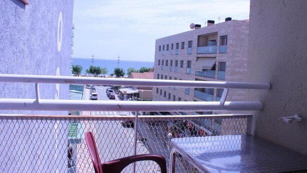 Un balcón con vista a una calle concurrida y edificios, una cerca de malla y una silla roja.