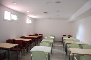 A classroom with rows of desks and chairs, white walls, and ceiling lights.