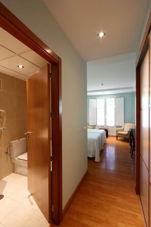 Ein Hotelkorridor mit einer Holztür, die zu einem Badezimmer führt, einem Bett im angrenzenden Raum und einem Sessel.