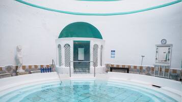 Una piscina interior con dos personas recostadas en el borde, una pared blanca con un arco verde y un reloj en la pared.