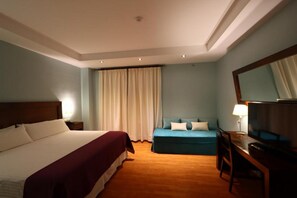 Ein Hotelzimmer mit einem großen Bett, einem kleinen Sofa, einem Schreibtisch mit Fernseher und einem Fenster mit Vorhängen.