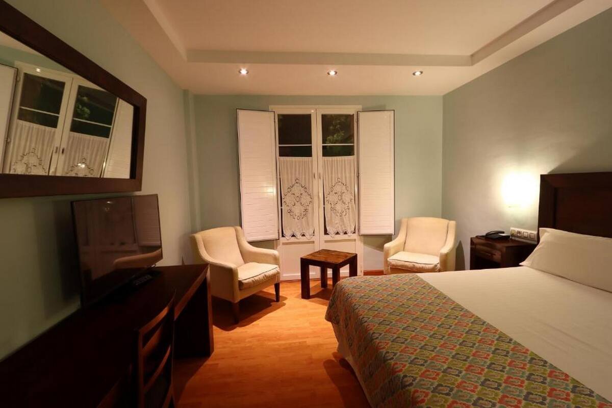 A hotel room with a bed, two chairs, a small table, a TV, and a window with curtains.