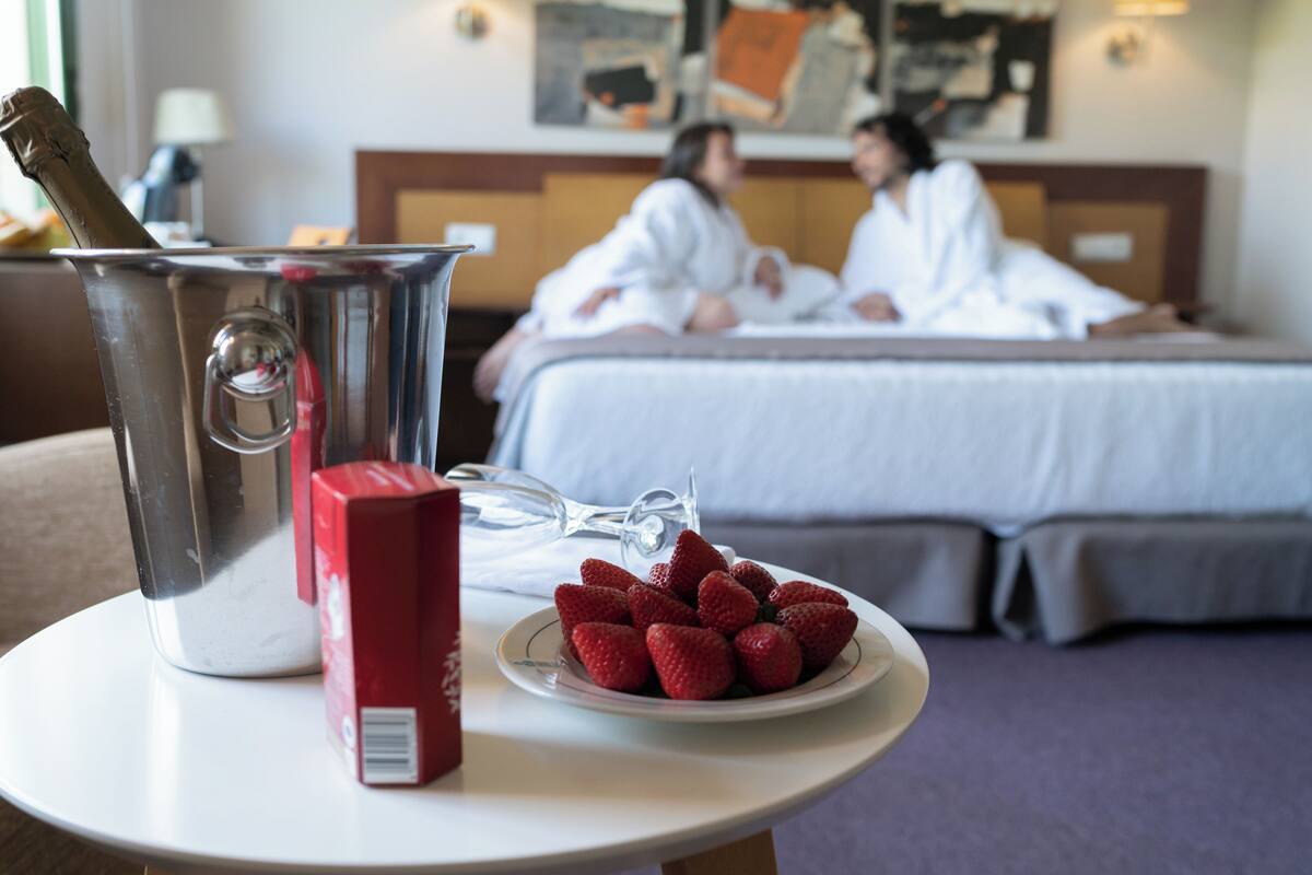 A hotel room with a bed, a bedside table with a champagne bottle, a red box, and a plate of strawberries.