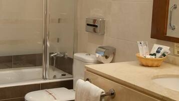 A bathroom with a glass shower enclosure, a bathtub, two toilets, a sink with a mirror, and a basket of toiletries.