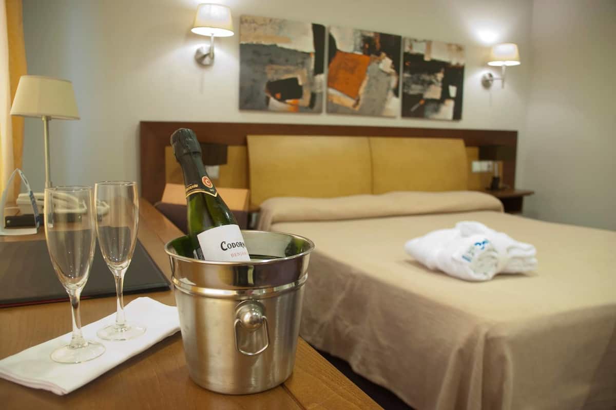 A hotel room with a bed, a nightstand, a lamp, a bottle of champagne, and two glasses on a table.