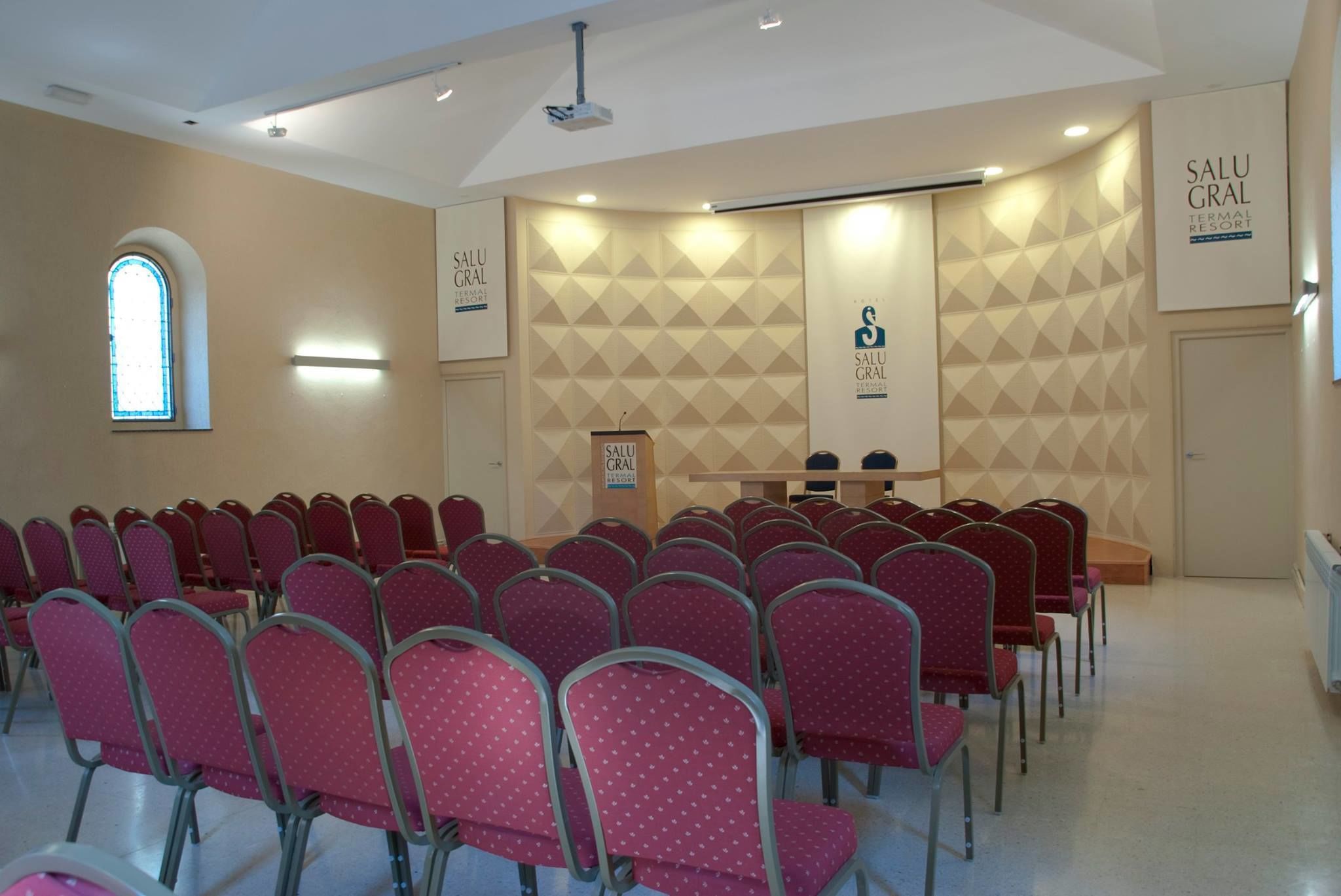 Un salón de conferencias con filas de sillas rojas, un atril y una pared con un diseño geométrico.