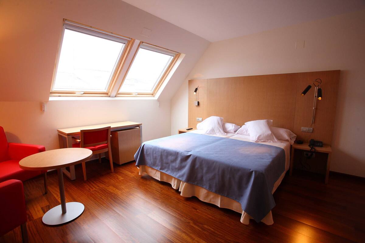 A room with a bed, a desk, a chair, and a small table. There are skylights above the bed.