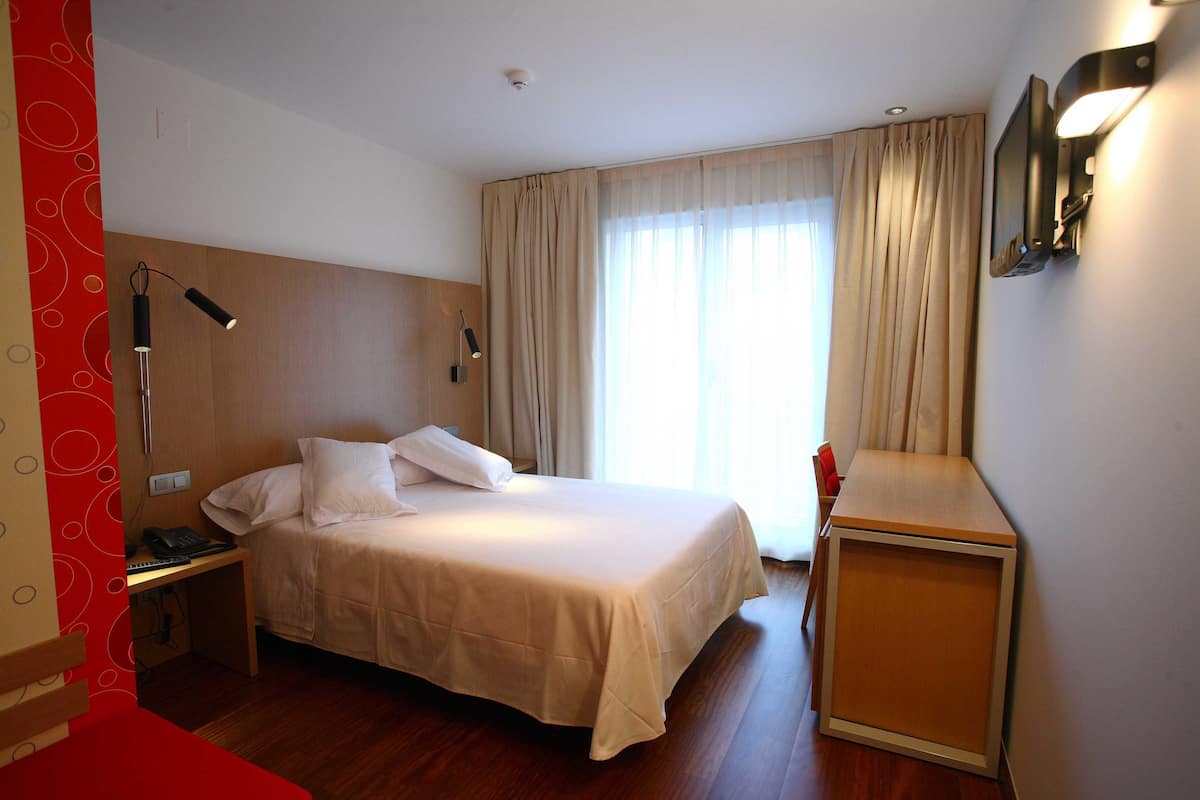 A hotel room with a bed, bedside table, lamp, and a small wooden side table.