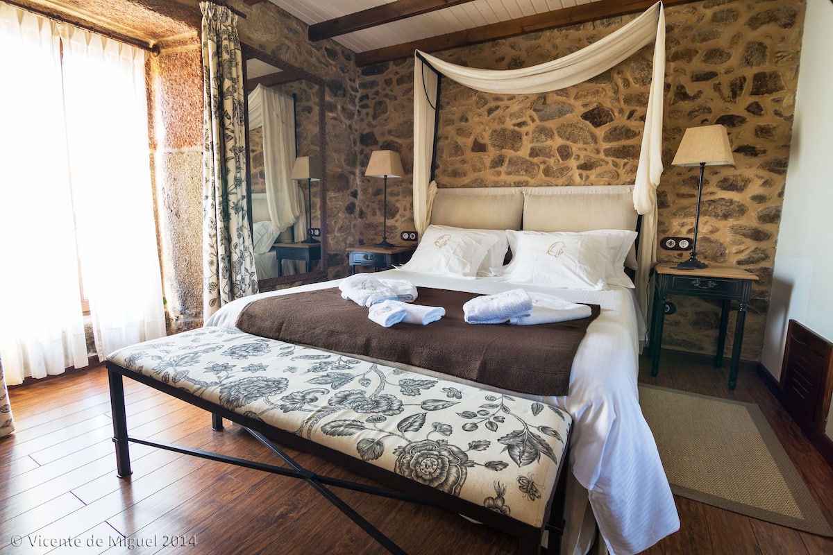 A four-poster bed with a canopy, a bedside table with a lamp, and a stone wall.