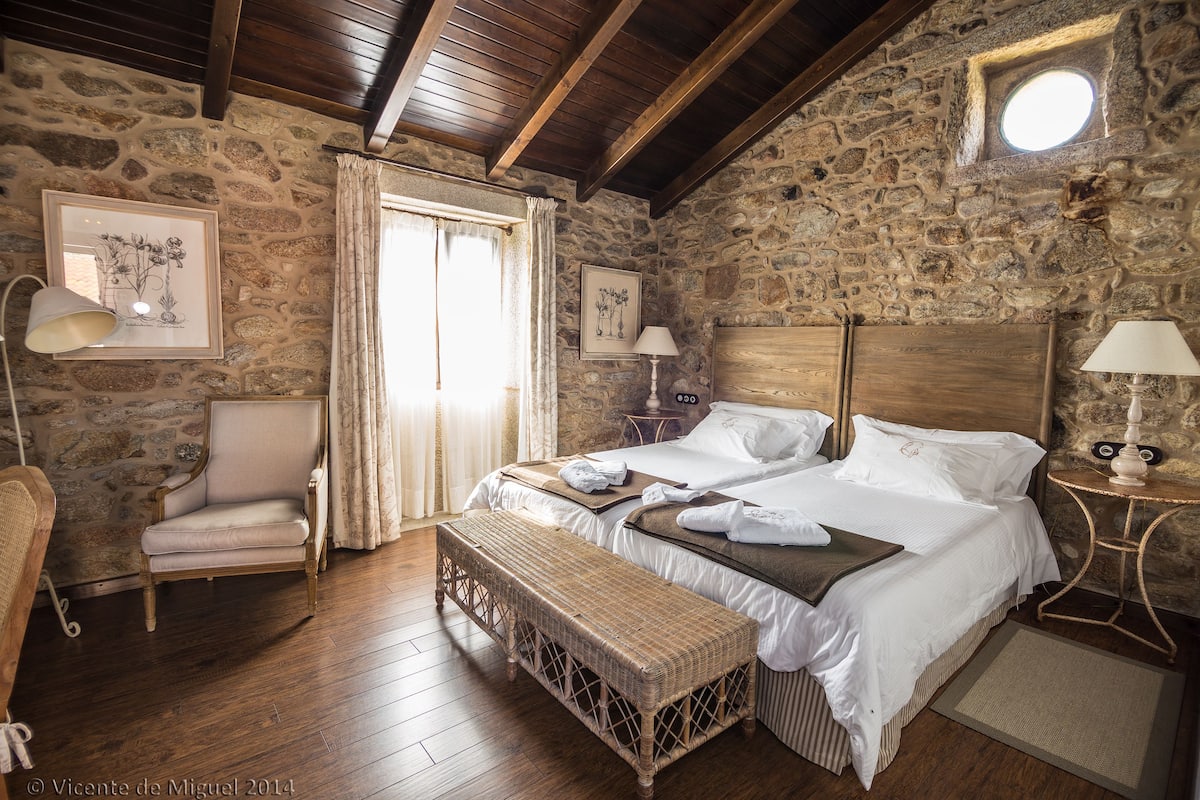 A rustic bedroom with stone walls, wooden floors, a large bed with white linens, a bench, a chair, and two framed pictures on the wall.