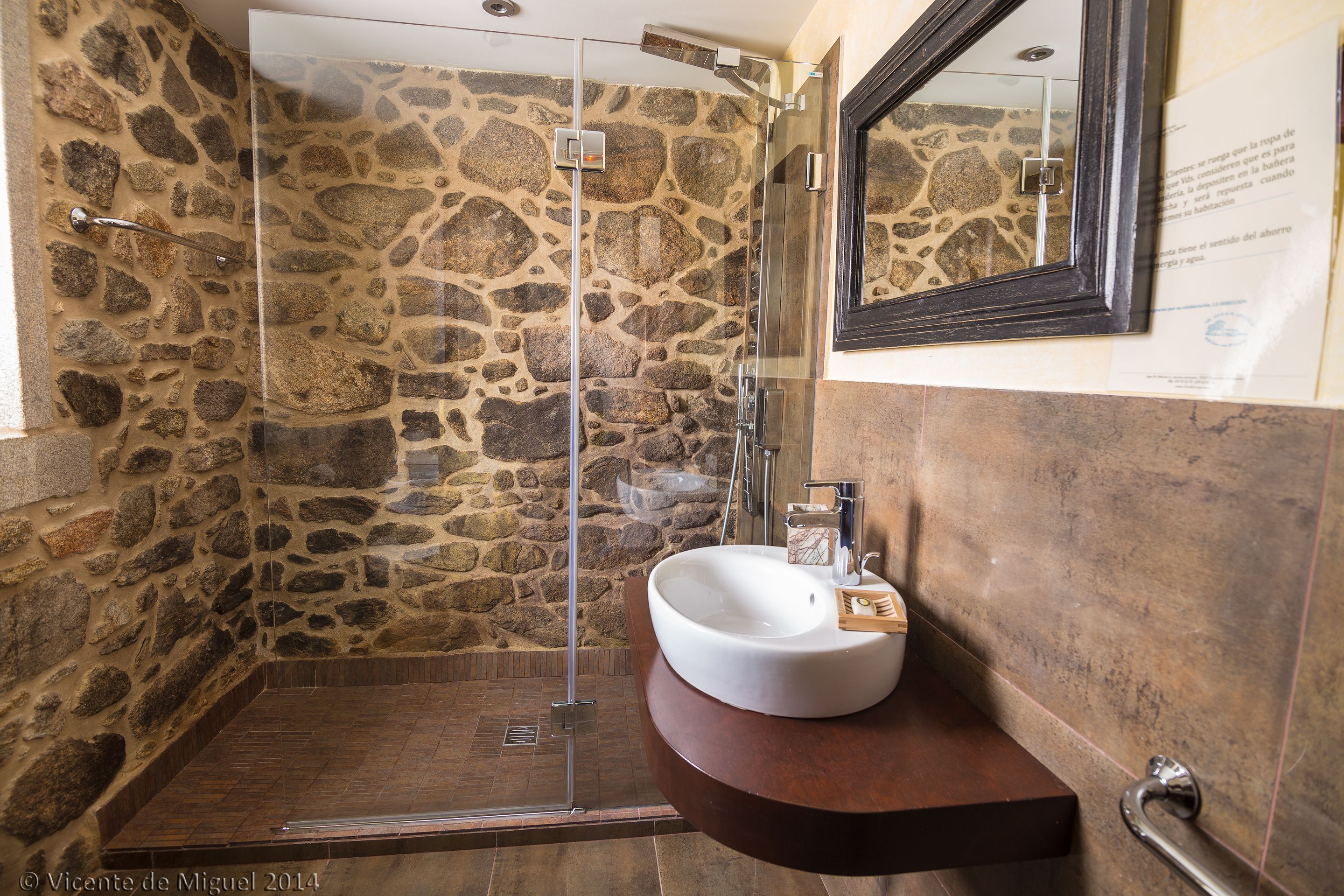 Un baño con pared de piedra, cabina de ducha de vidrio y lavamanos blanco con encimera de madera.