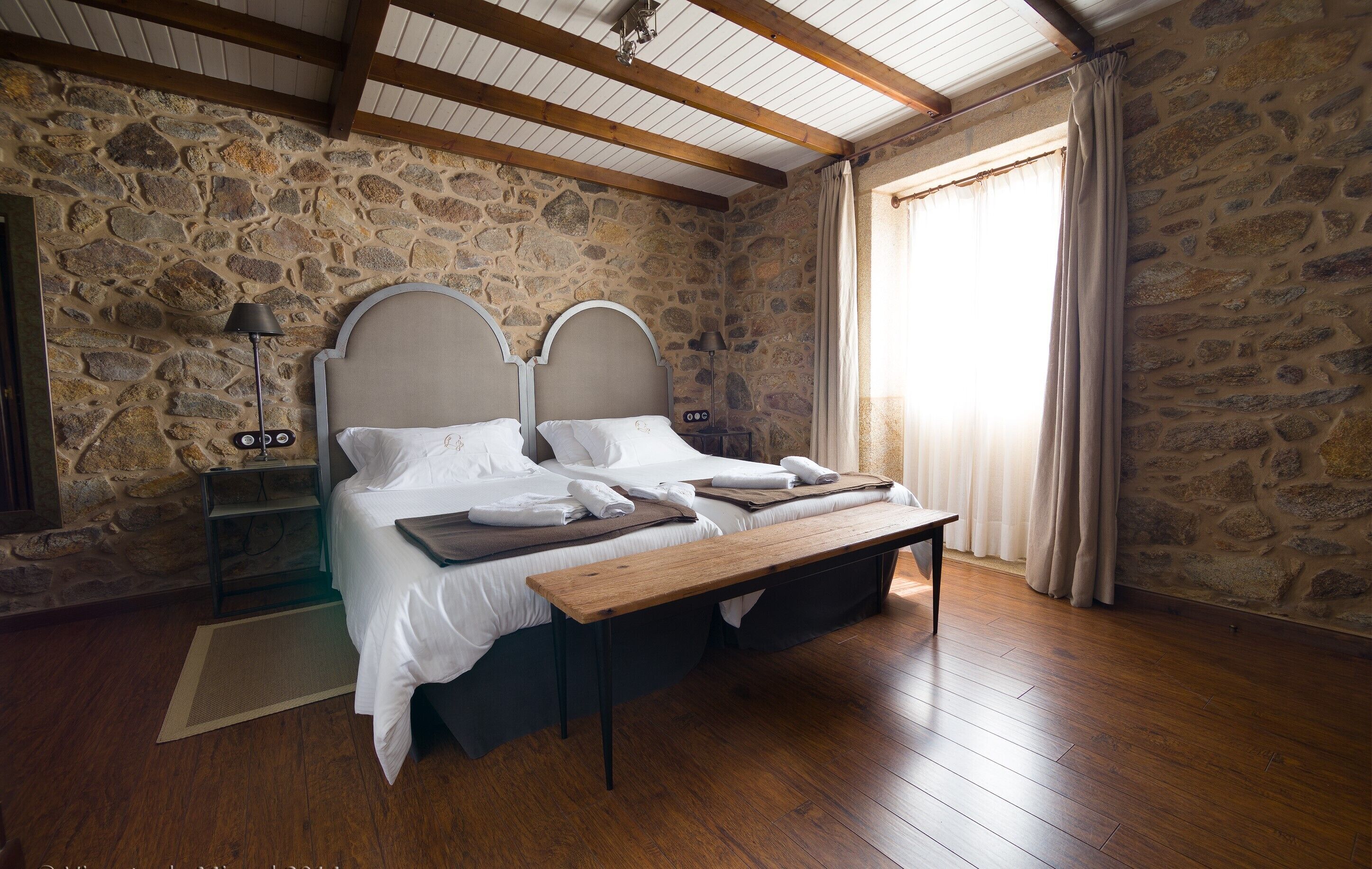 A bedroom with a stone wall, wooden beams, a bed with white linens, a bedside table, and a nightstand.