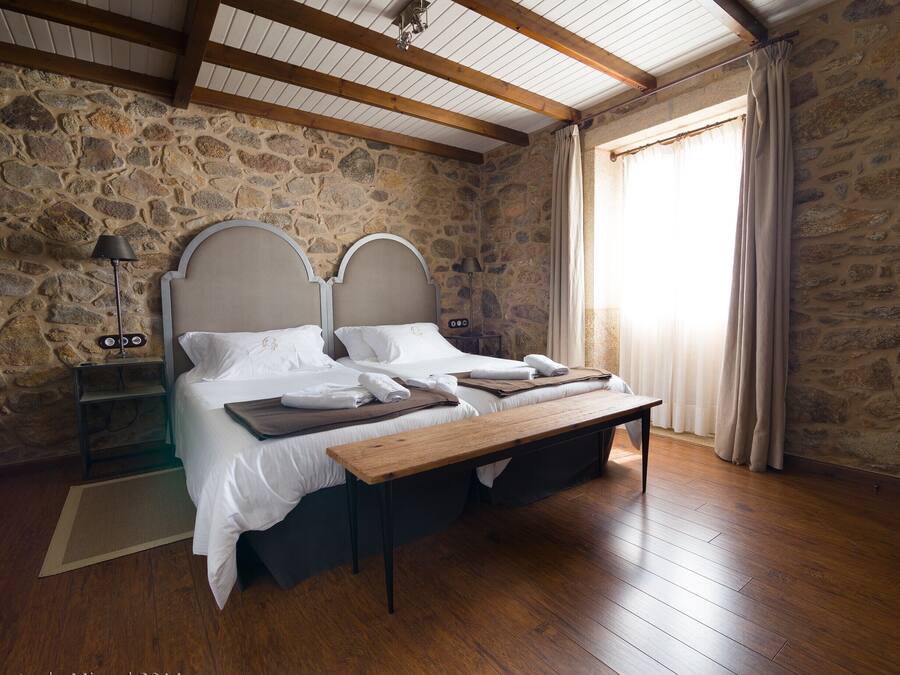 A bedroom with a stone wall, wooden beams, a bed with white linens, a bedside table, and a nightstand.