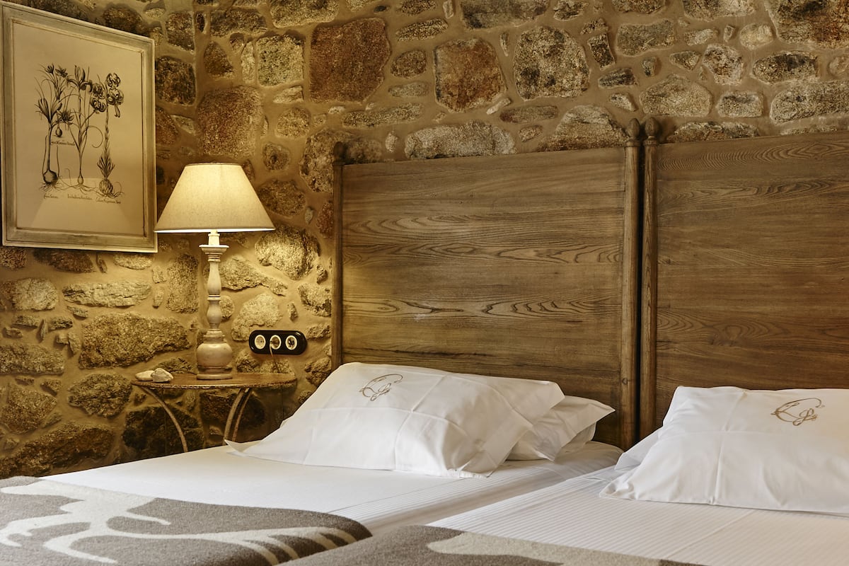 A double bed with a wooden headboard, a bedside table with a lamp, and a stone wall.